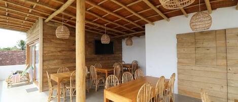 An outdoor dining area with wooden tables and chairs, woven hanging lights, and a TV mounted on the wall.