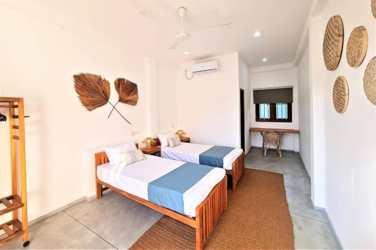 Quarto com duas camas, ventoinha no teto, ar condicionado e uma escrivaninha com cadeira.