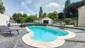 A swimming pool with a curved edge, surrounded by a tiled deck. There are lounge chairs and a small building in the background. - Charmante maison avec piscine (Le Château-d'Oléron)