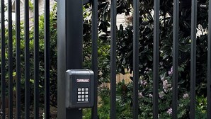 A metal gate with a keypad lock, surrounded by greenery and a stone wall.