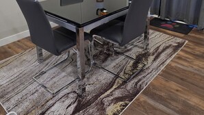 Un comedor con una mesa de vidrio, cuatro sillas, un florero con flores, un cuadro en la pared y una cortina.