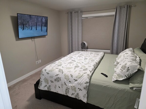 A bedroom with a bed, a TV mounted on the wall, a window with curtains, and a fan. - Grand Stylish Home | TVs in Bedrm & Lvroom | Comfy mattresses |Garage park | BBQ (Regina)