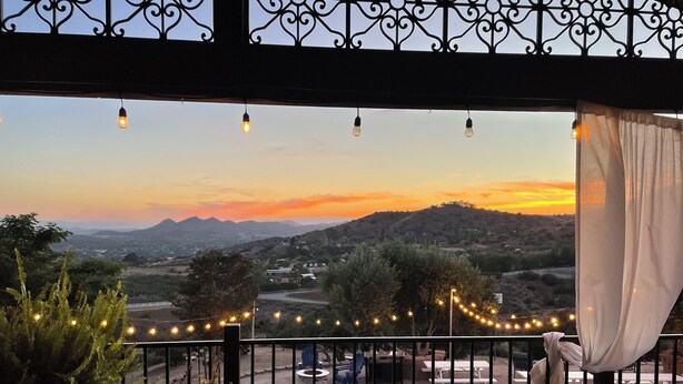 Un balcón con vistas a un atardecer sobre un paisaje montañoso, luces colgantes y mobiliario exterior.