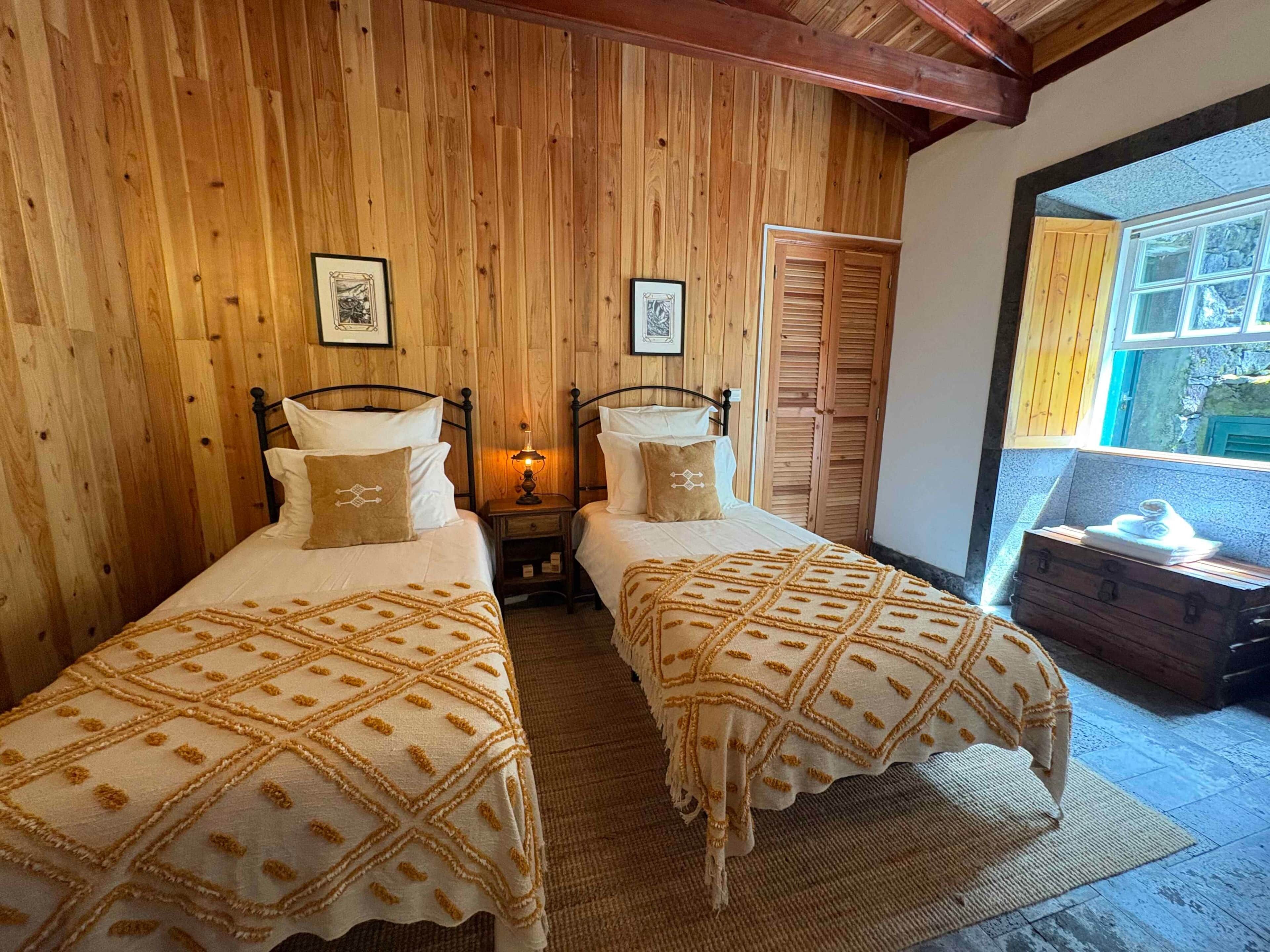 Une chambre avec deux lits, des murs en bois, une fenêtre donnant sur un beau paysage et une petite table sur laquelle se trouve une valise.