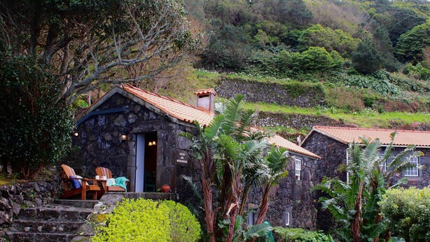 Un edificio de piedra con tejado de tejas, una escalera que lleva a un área de estar con sillas, y exuberante follaje rodeando la propiedad.