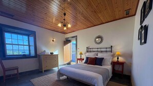 A bedroom with a wooden ceiling, a large window, a bed with white bedding, two bedside tables with lamps, a wooden dresser, and a wall clock. - Casa do Jardim (Santo Amaro)