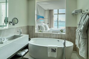 A modern bathroom with a large bathtub, a vanity with a sink, a mirror, and a window with a city view. - Th8 Palm Dubai Beach Resort, Vignette Collection (Dubai)