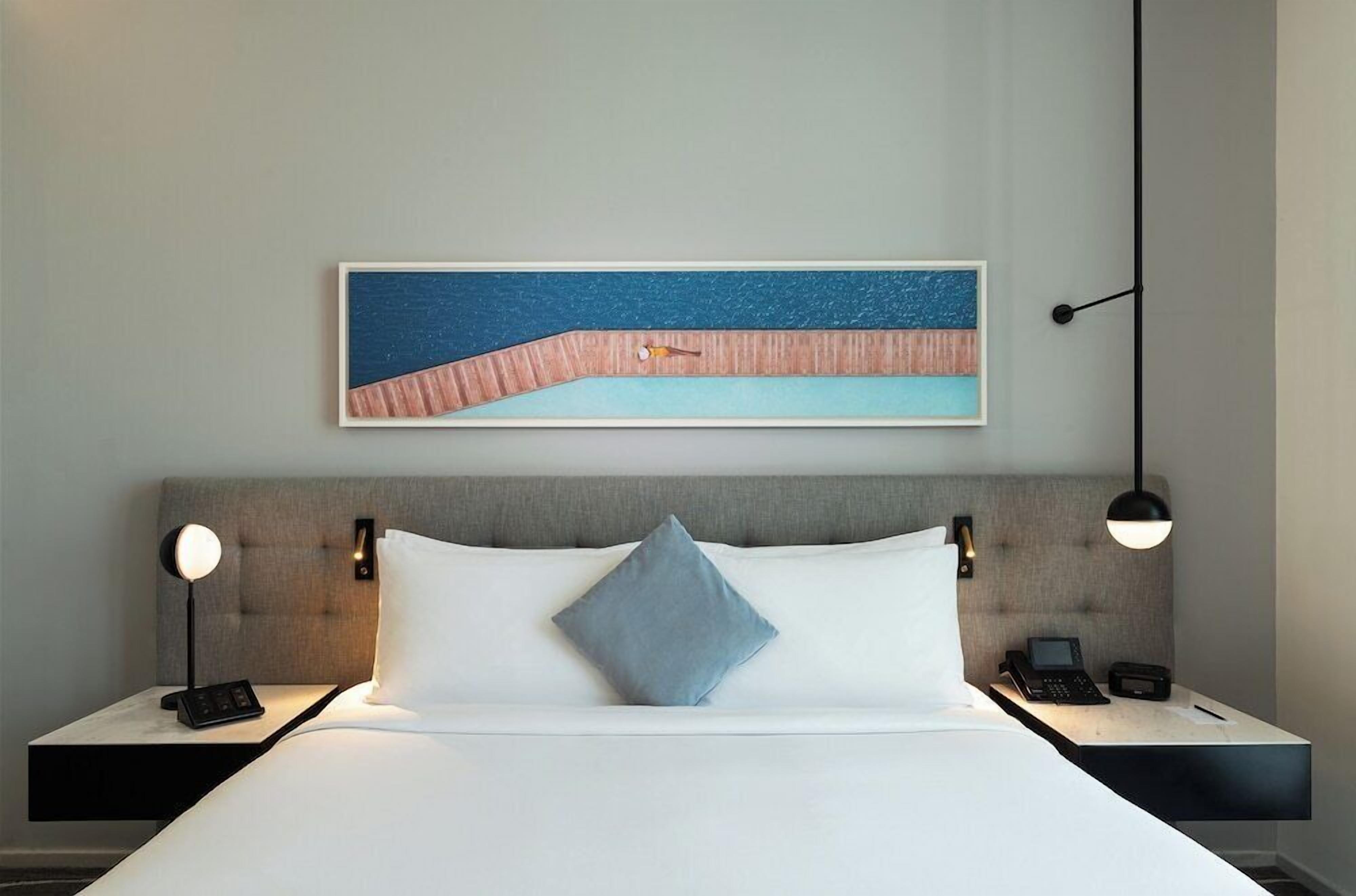 A hotel room with a bed, a nightstand with a phone, a lamp, and a painting on the wall.