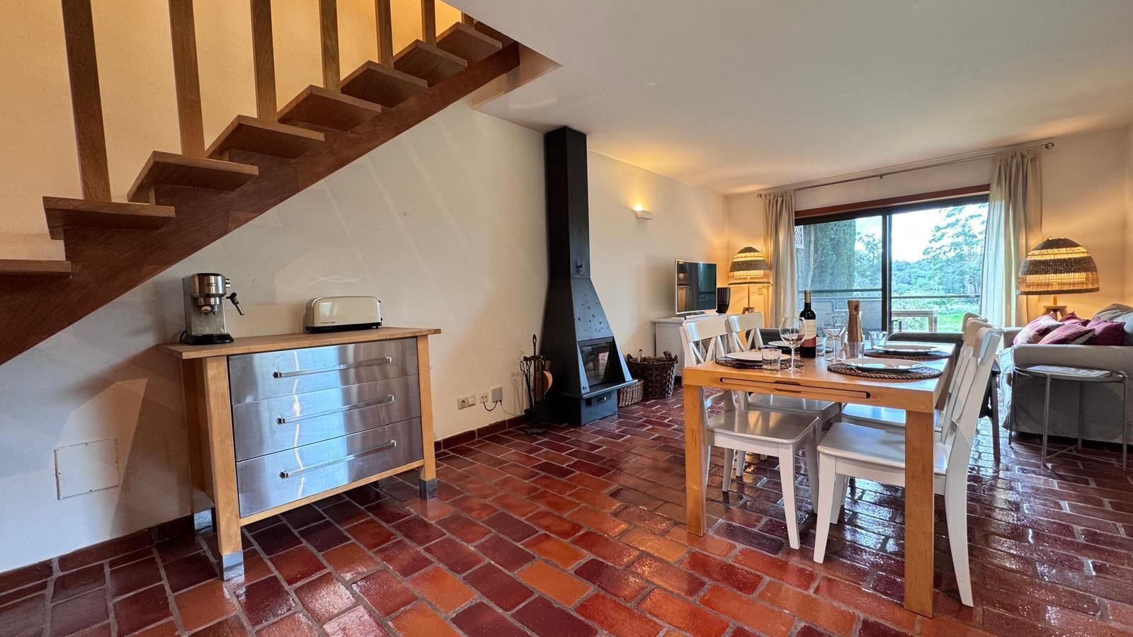 A kitchen with a wooden staircase, a dining area with a table and chairs, and a view of the outdoors through a window.