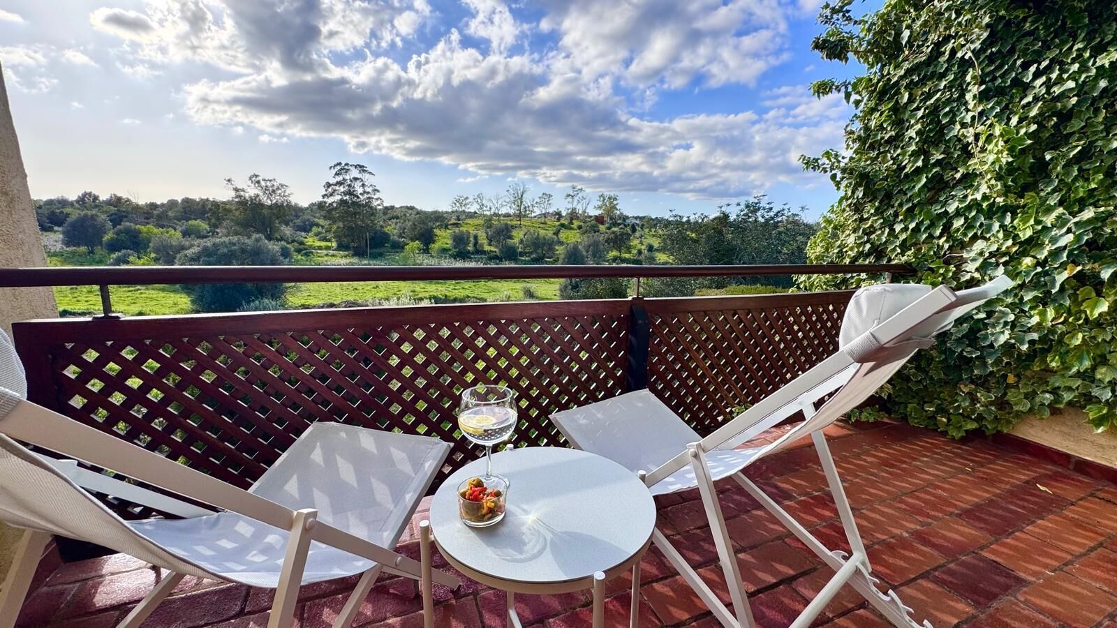 En balkon med udsigt over et frodigt landskab, to hvide stole, et lille rundt bord og et glas vin.