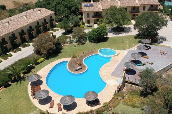 A swimming pool with a wooden deck, surrounded by lounge chairs and umbrellas, with buildings in the background.