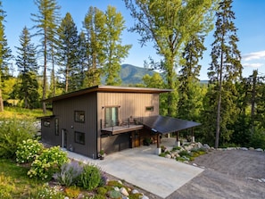 A modern two-story cabin with a balcony, surrounded by a natural landscape with trees and mountains. - New | 5 Min to Glacier | Hot Tub & Fire Pit (Martin City)