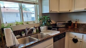 A kitchen with a granite countertop, stainless steel sink, white cabinets, a dishwasher, a toaster, and a window with plants. - Modern 1-bedroom apartment w/Pool in Popular Northpark Neighborhood (San Diego)