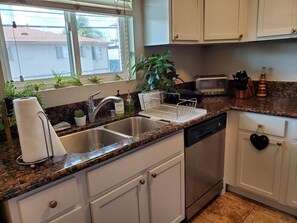 Eine Küche mit Granit-Arbeitsplatte, Edelstahl-Spüle, weißen Schränken, einem Geschirrspüler, einem Toaster und einem Fenster mit Pflanzen.