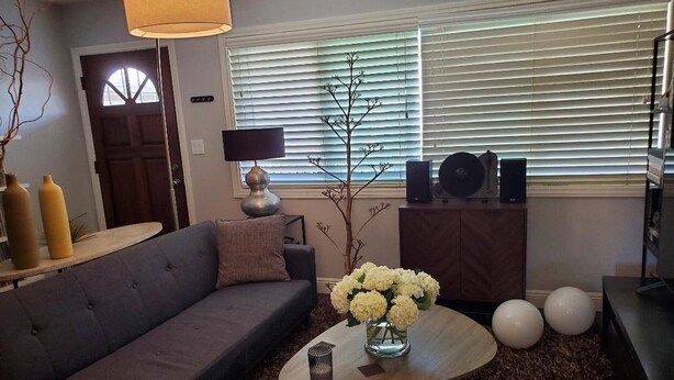 A living room with a sofa, a coffee table with a vase of flowers, a lamp, and a window with blinds. - Modern 1-bedroom apartment w/Pool in Popular Northpark Neighborhood (San Diego)