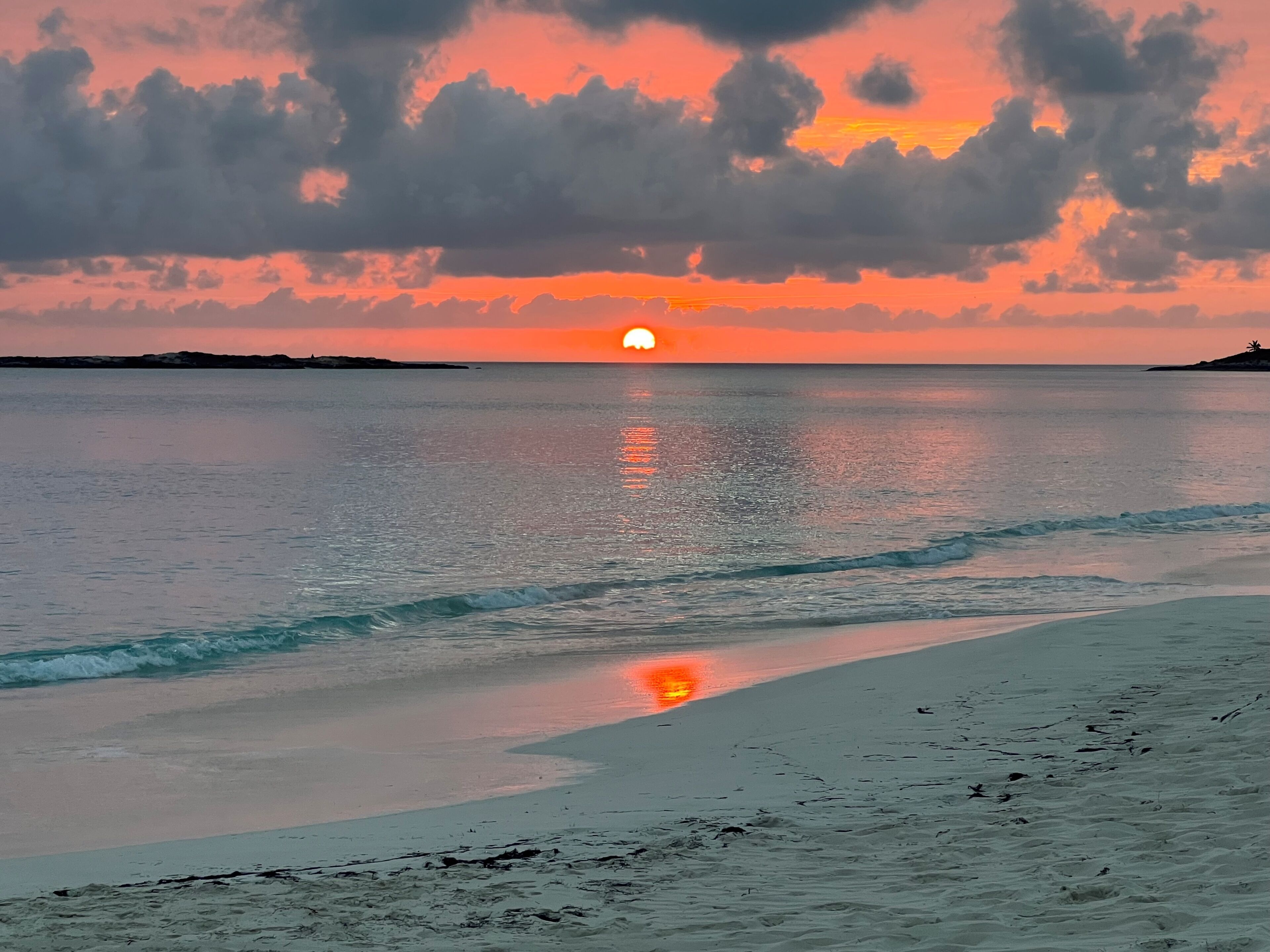 A beach with a sunset, calm water, and a distant landmass.