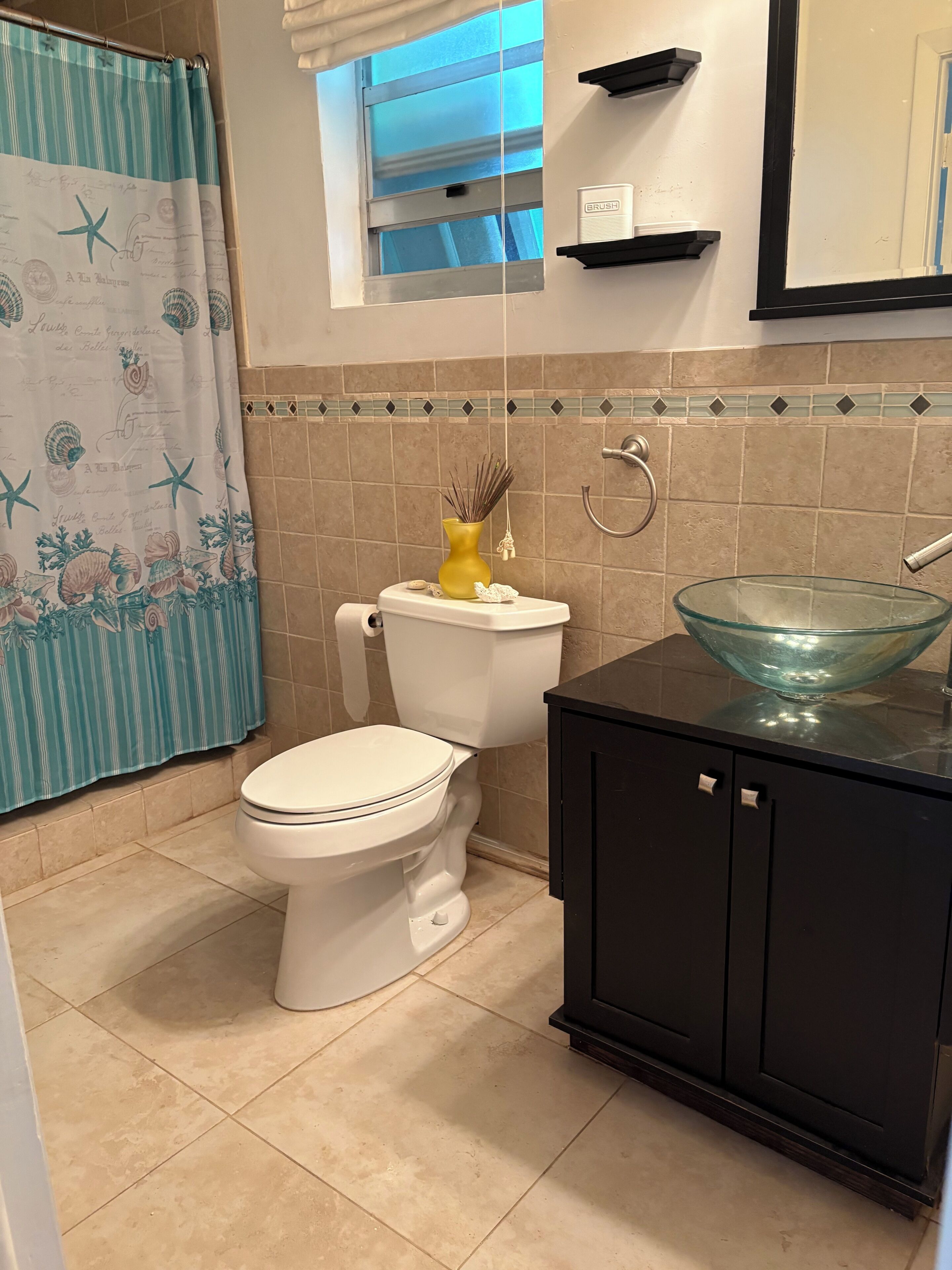 A bathroom with a toilet, a glass bowl sink, a black cabinet, a towel rack, and a shower curtain with a nautical theme.