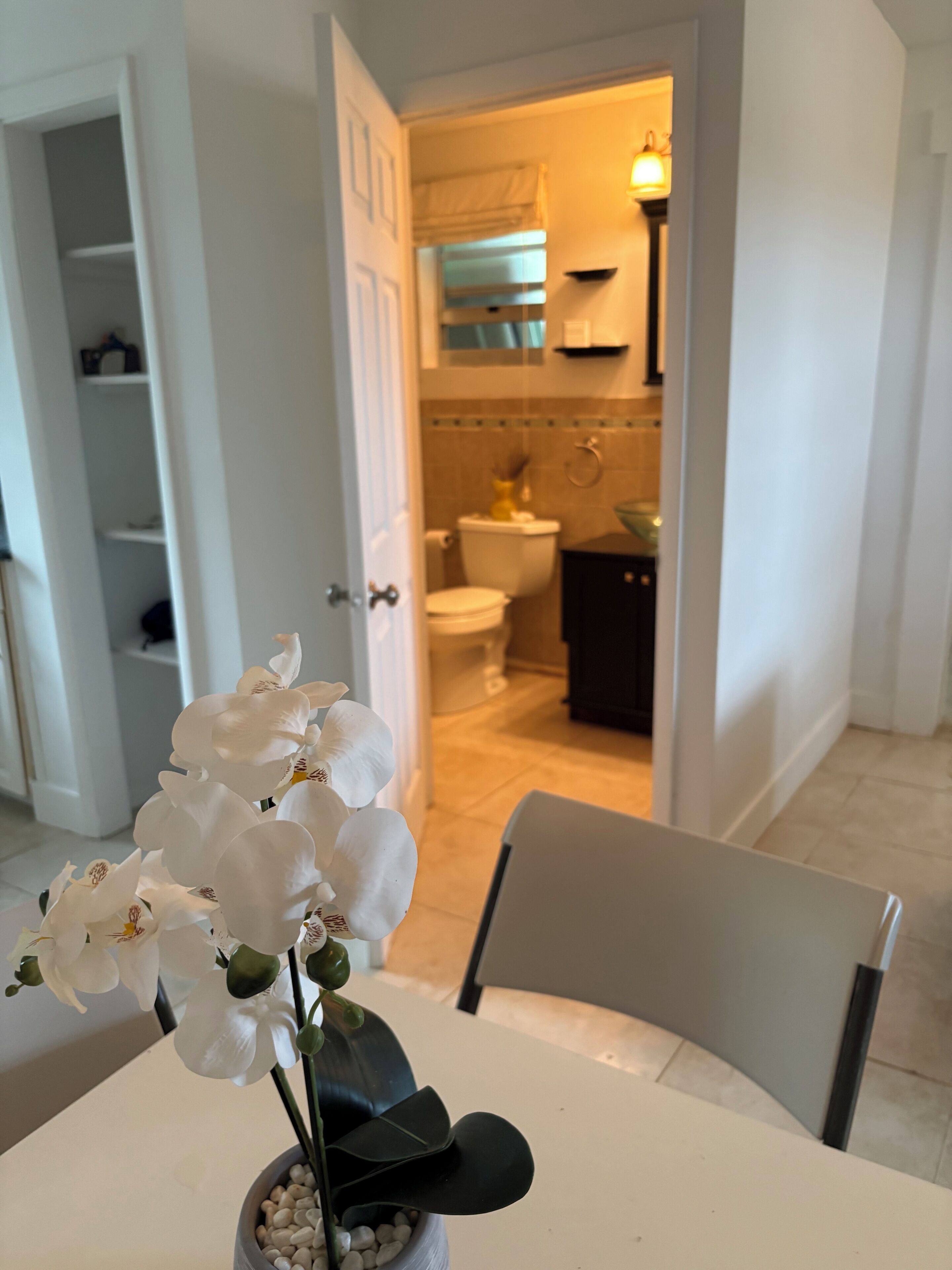 A white dining table with a vase of white orchids, a chair, and a view into a bathroom with a toilet, sink, and towel rack.