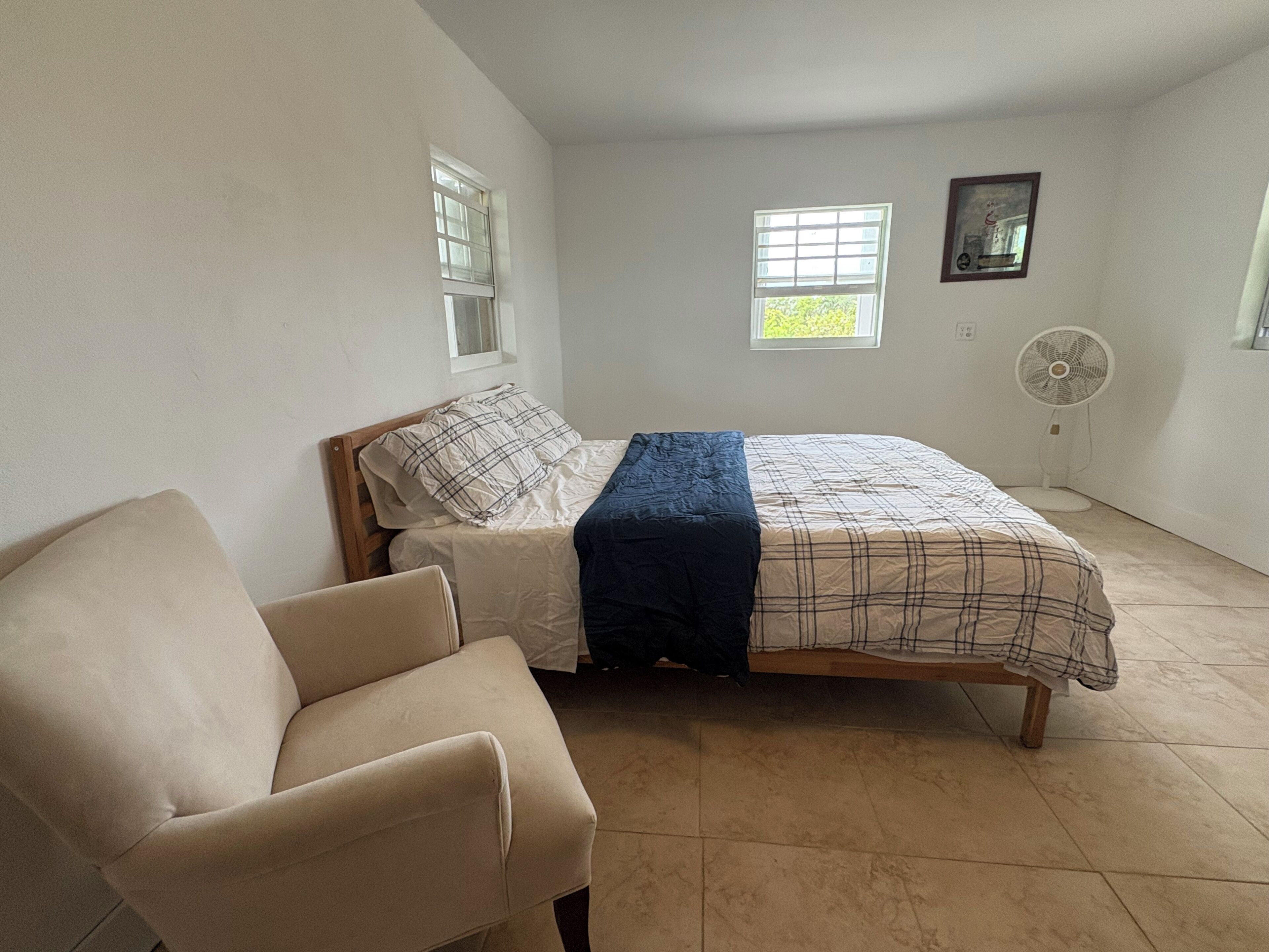 Une chambre à coucher avec un lit, une chaise, deux fenêtres, un ventilateur et un tableau accroché au mur.
