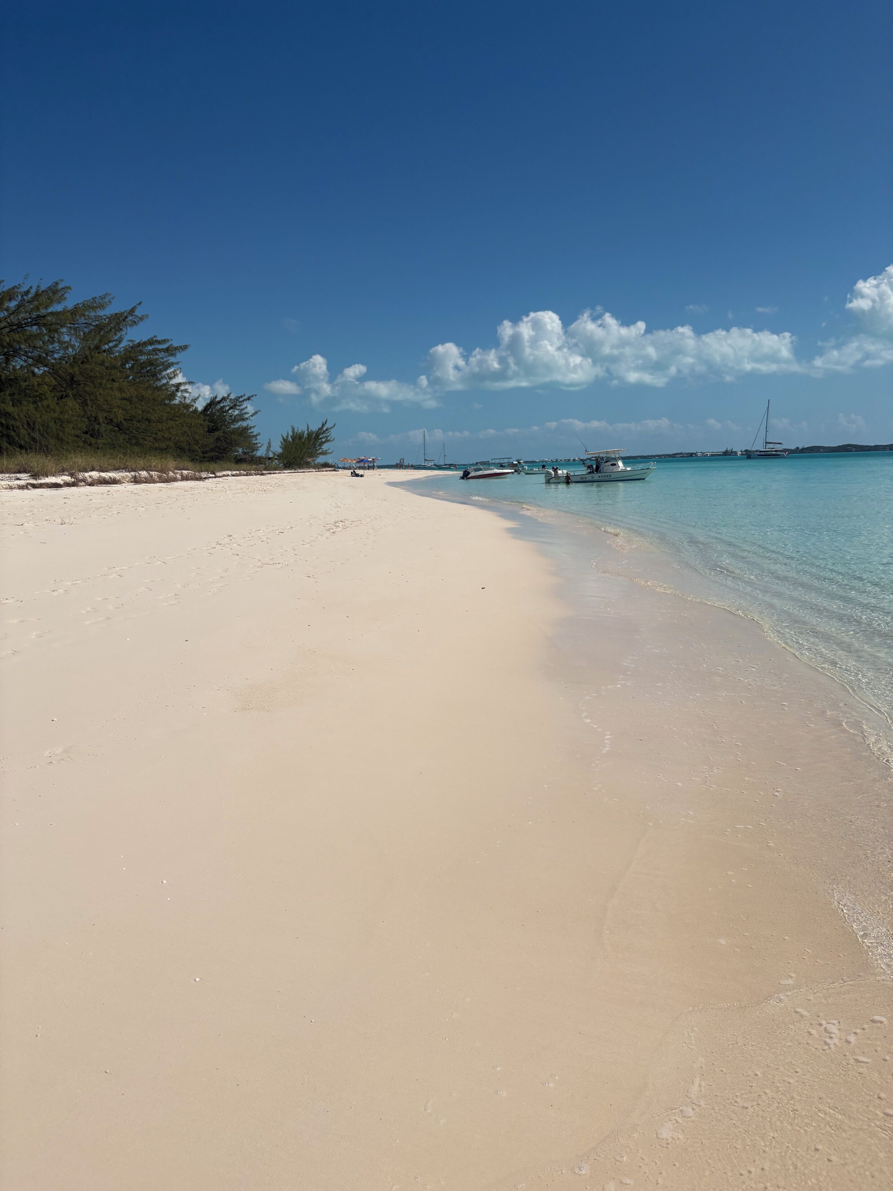 A beach with clear turquoise water, a sandy shore, and boats anchored in the distance.