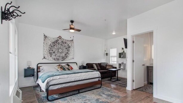 Un dormitorio con cama, ventilador de techo, tapiz, mesita de noche, cómoda y una puerta que da a otra habitación.