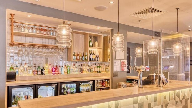 Een bar met een toonbank, planken vol flessen en hangende lampen.