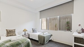 A bedroom with two beds, a bedside table with a lamp, a window with blinds, and a small table with a vase. - The Coral Esplanade with Pool (Mornington)