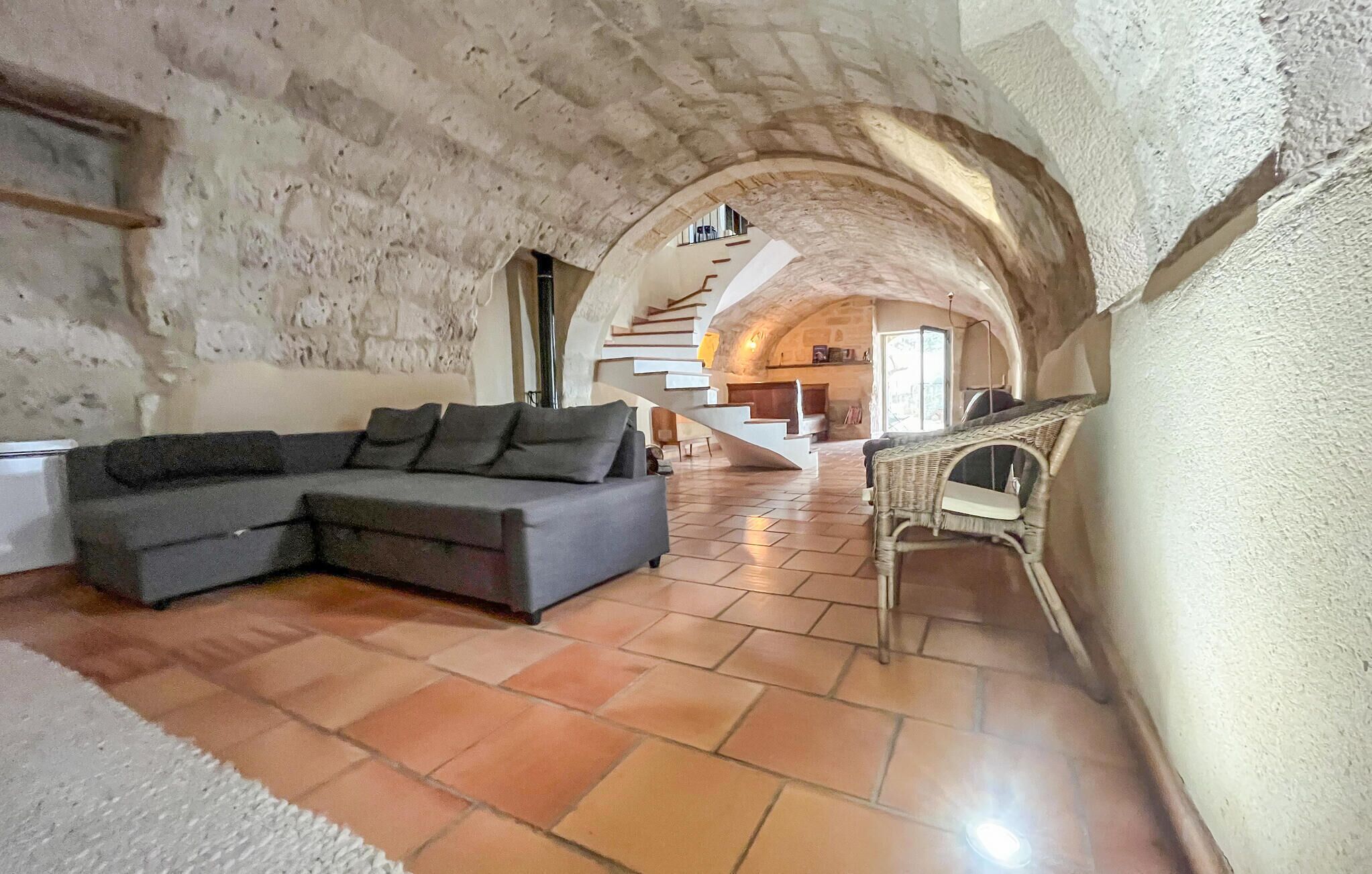Ein Innenraum mit einer Steinwand, einem grauen Ecksofa, einem Korbstuhl und einer Treppe, die zu einer Erhöhung führt.