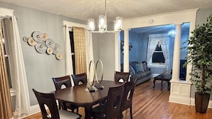 A dining area with a wooden table and chairs, a chandelier, and a view into a living room with a blue wall and a plant.