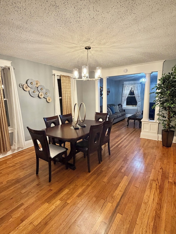 A dining area with a wooden table and chairs, a chandelier, and a view into a living room with a blue wall and a plant.