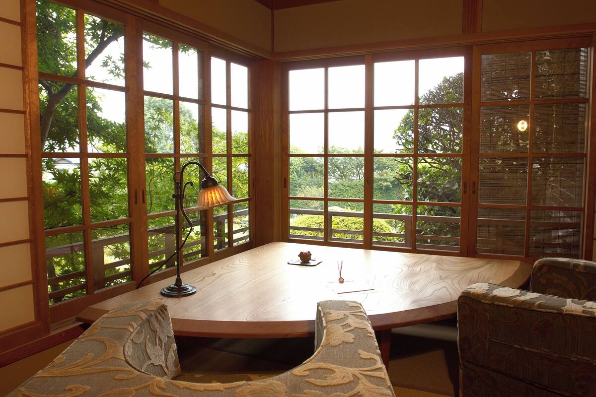 A room with a round table, two chairs, a lamp, and large windows with a view of greenery.