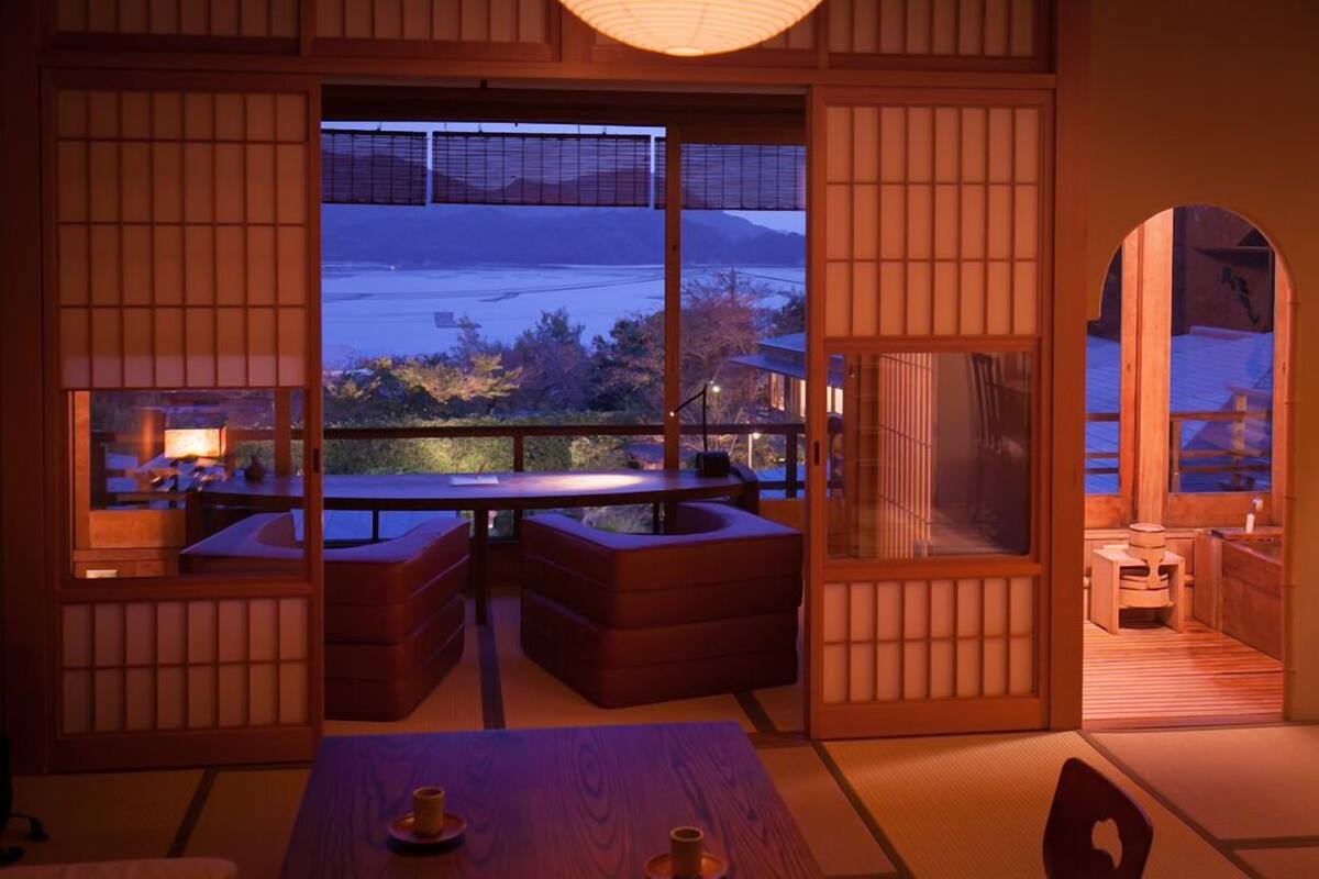 A traditional Japanese room with tatami flooring, a low table, cushions, and a view of a body of water.