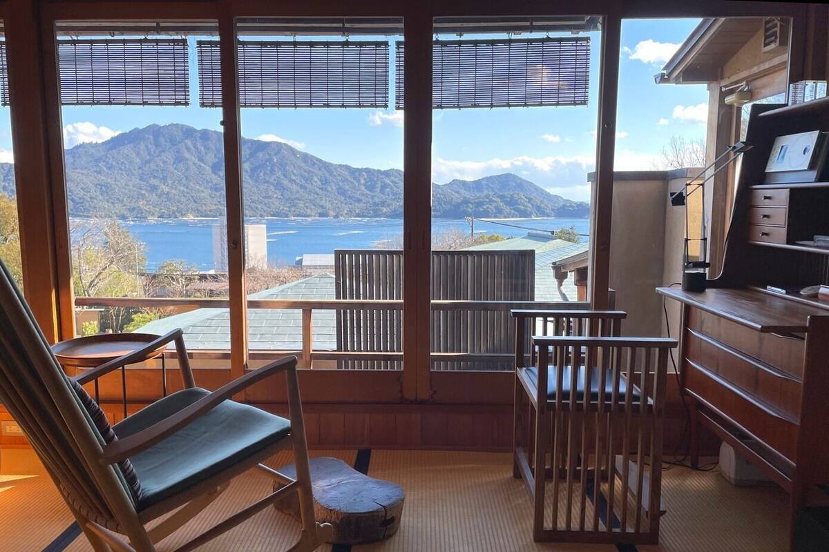 A room with a view of mountains and a body of water, a wooden desk, a chair, and a tatami mat floor.