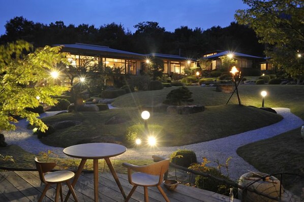 A well-lit outdoor area with a round table and chairs, surrounded by a landscaped garden with pathways and illuminated lights. - Miyahama Onsen Teien no Yado Sekitei (Hatsukaichi)