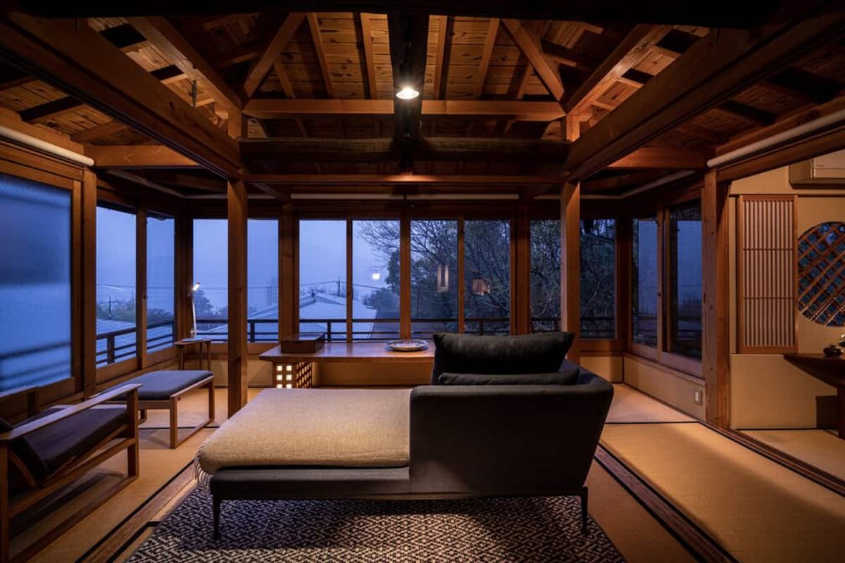 A traditional Japanese-style room with tatami flooring, a low wooden ceiling, and large windows offering a view of the outside.