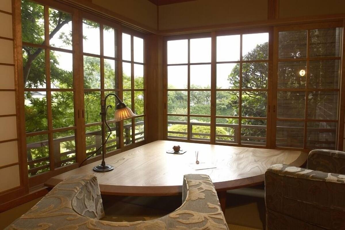 A room with a round table, two chairs, a lamp, and large windows with a view of greenery.