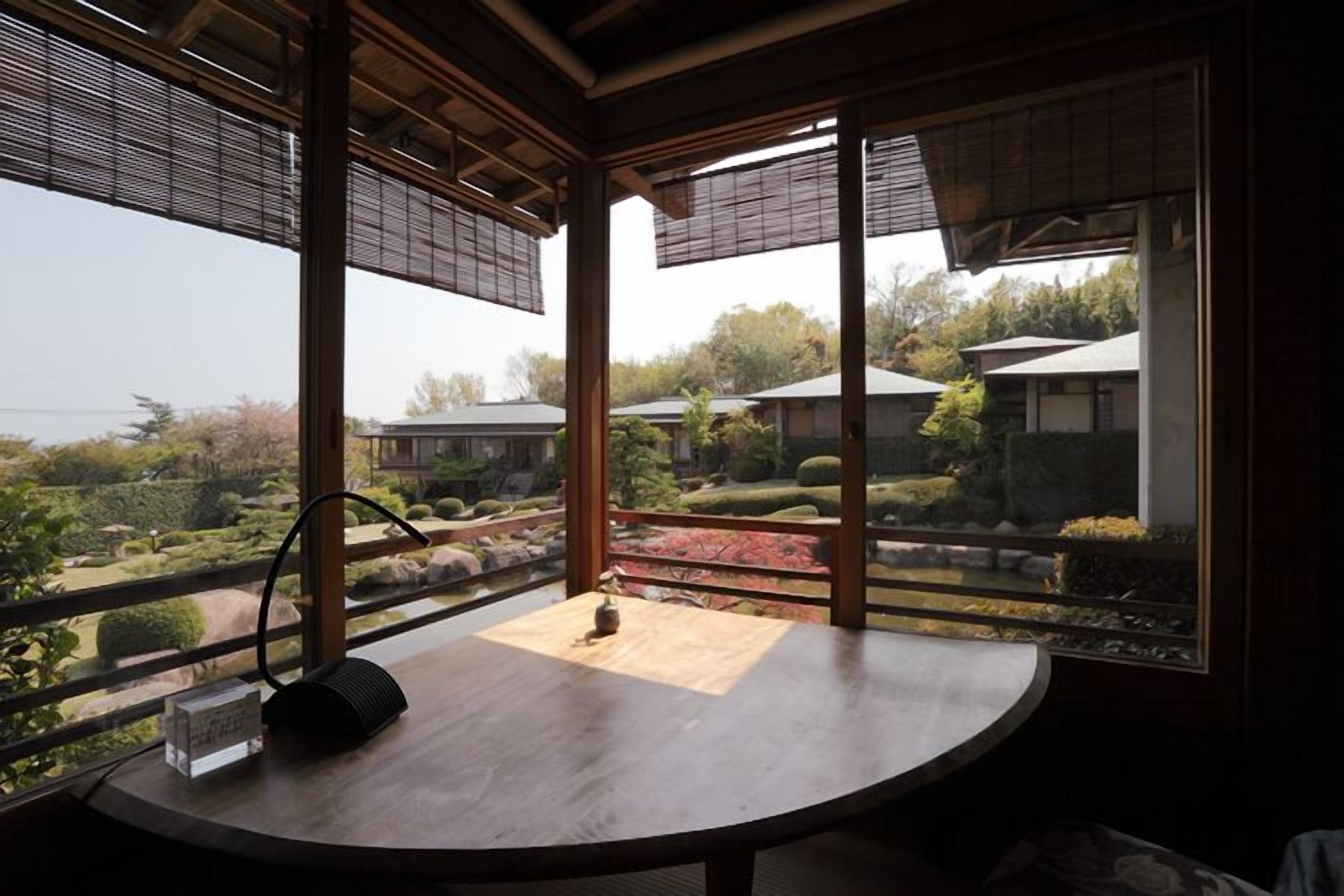 A room with a wooden table, a clock, and a view of a garden and buildings outside.