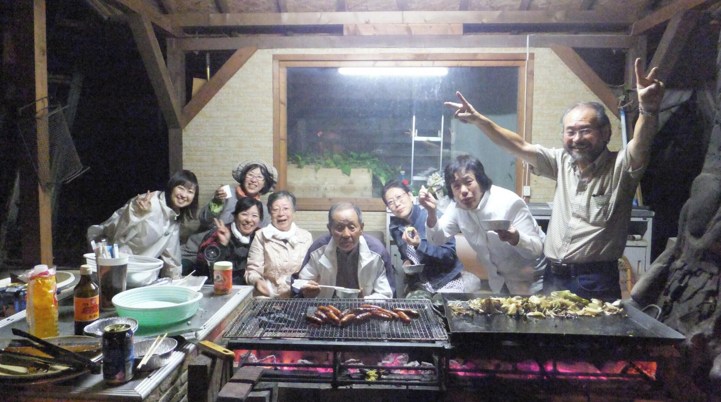 Un grupo de personas reunidas alrededor de una parrilla con salchichas, en una habitación con paredes de ladrillo y un ventanal grande.