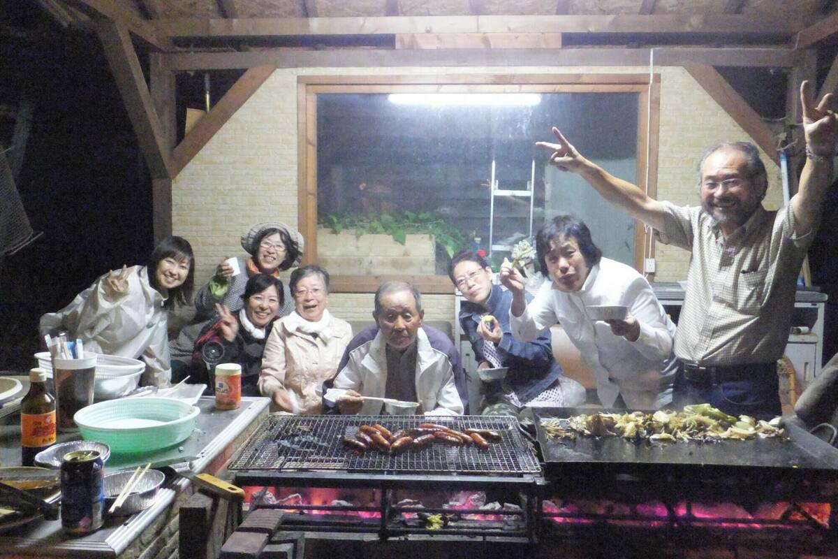 A group of people gathered around a grill with sausages, in a room with brick walls and a large window.