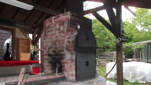 A rustic outdoor kitchen with a brick oven, wooden logs, and a covered seating area. - Hitomoshi no Sato (Nanae)