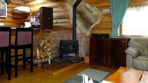 A cozy cabin interior with a fireplace, wooden walls, a dining area, and a leather couch. - Hitomoshi no Sato (Nanae)