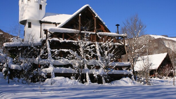 A snow-covered wooden cabin with a large chimney, surrounded by snow-laden trees and a clear blue sky. - Hitomoshi no Sato (Nanae)