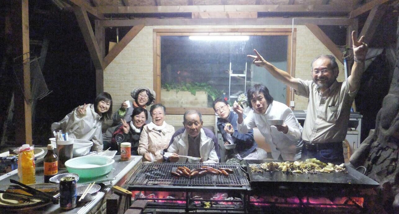 A group of people gathered around a grill with food, in a room with wooden beams and a window.