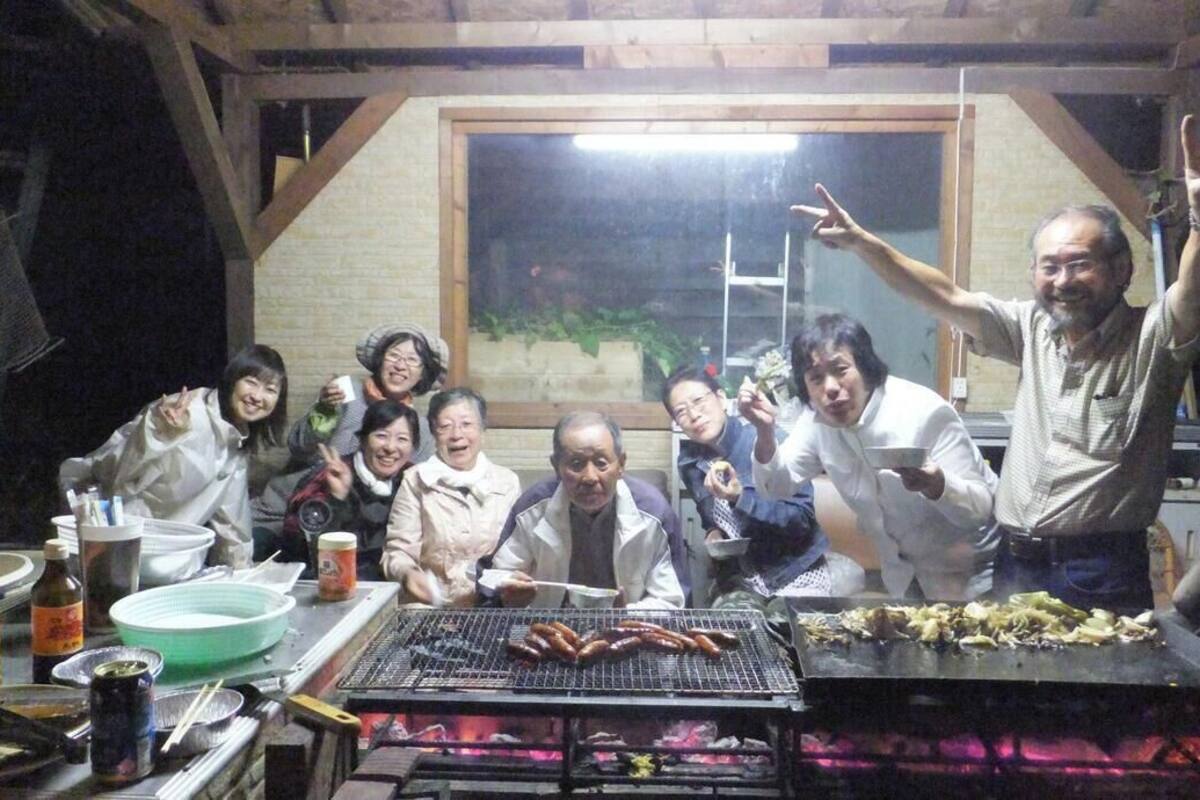 A group of people gathered around a grill with food, in a room with wooden beams and a window.