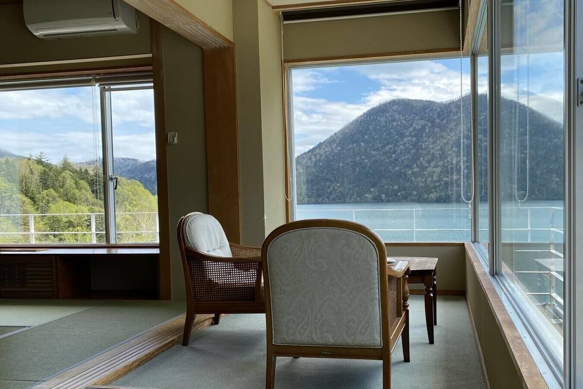 Una habitación con vista a un lago y montañas, una silla de madera, una mesita y un ventanal grande.