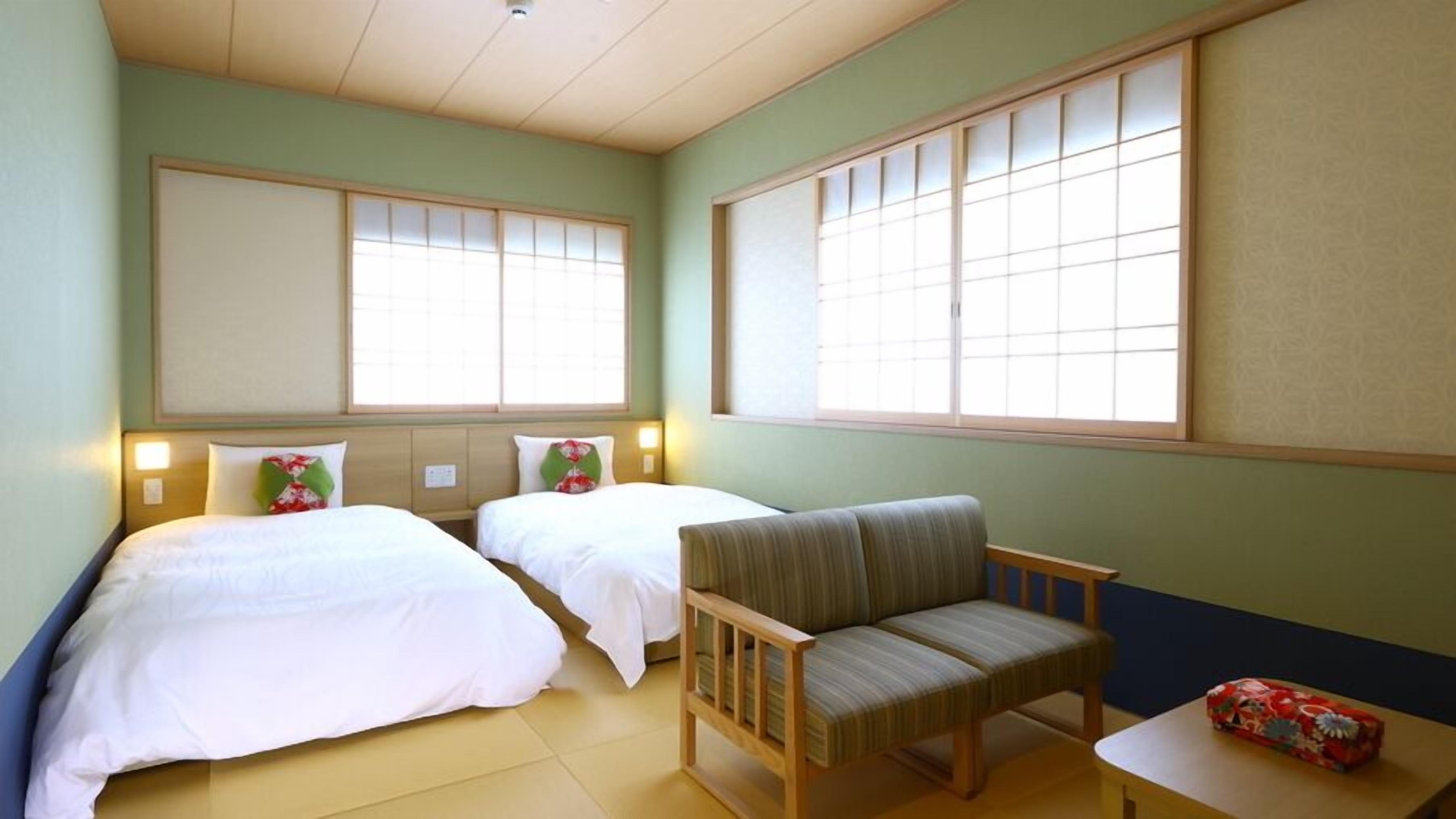Ein Hotelzimmer mit zwei Betten, einem Sessel, einem kleinen Tisch und großen Fenstern.