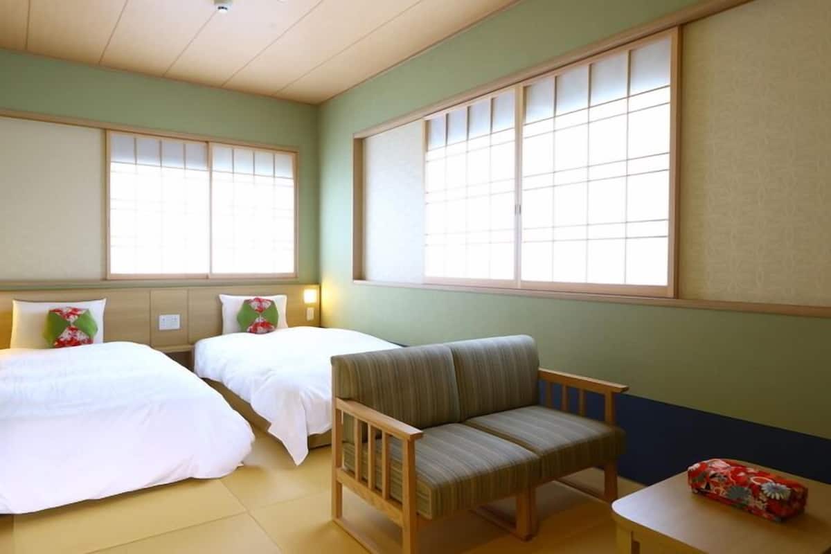 A hotel room with two beds, a chair, a small table, and large windows.