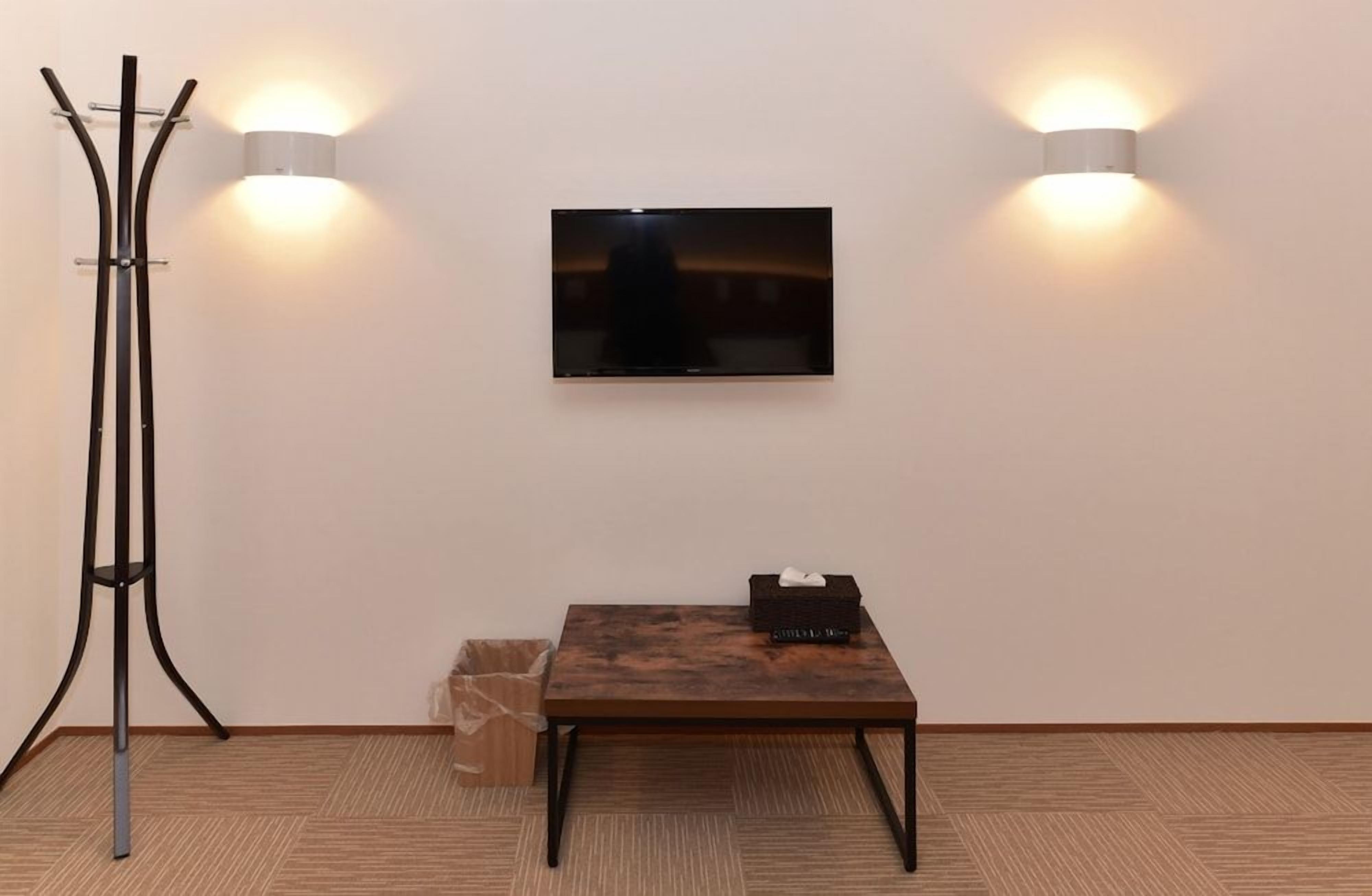 A room with a flat-screen TV mounted on the wall, a wooden table with a black box on it, and a tall floor lamp.