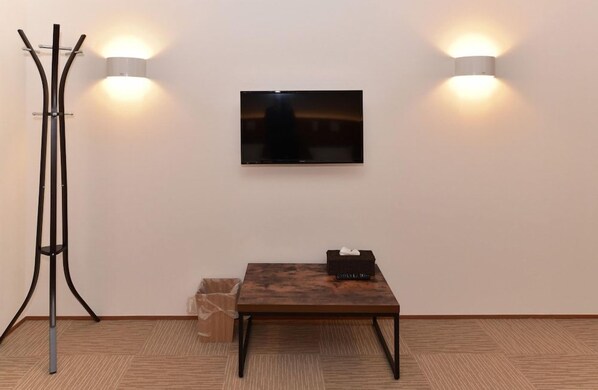 A room with a flat-screen TV mounted on the wall, a wooden table with a black box on it, and a tall floor lamp. - Coco Shuku Nakano (Tokyo)