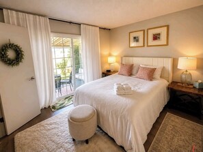 A bedroom with a bed, two bedside lamps, a nightstand, a round ottoman, a window with curtains, and a wall-mounted wreath. - Beautiful 2-bedroom building in charming Sacramento (Sacramento)